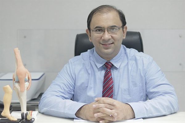 Dr. Kuldeep Gadkari, smiling, at his clinic desk beside an anatomical model of the knee